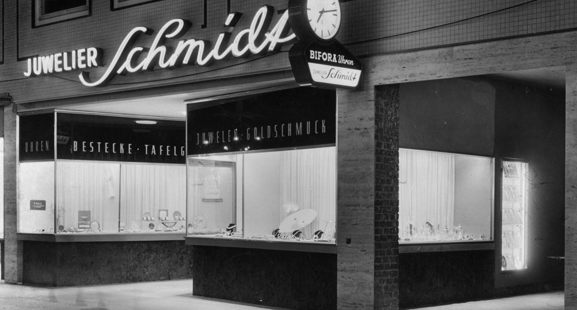 Schaufenster von Juwelier Schmidt bei Nacht mit Leuchtschrift und Bifora-Uhr