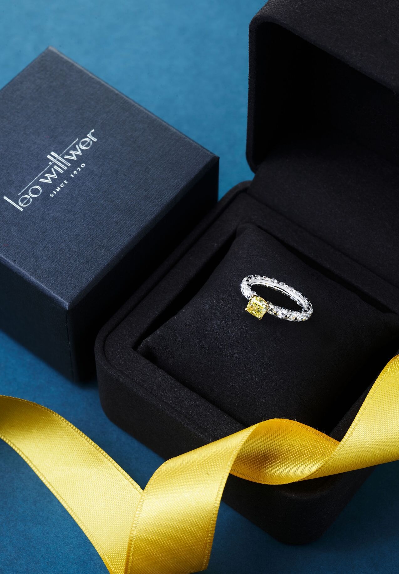 Verlobungsring mit gelbem Diamanten in einem offenen Leo-Wittwer-Etui, daneben ein gelbes Satinband